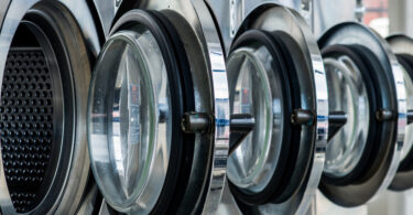 Four steel front-loaded laundry machines in a laundry facility with their round doors swung open.